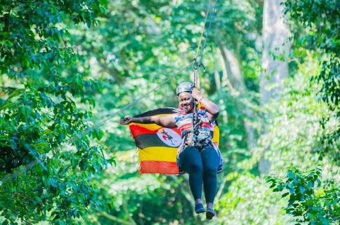 Tourist enjoying a zipline adventure in Mabira Forest, Uganda, while holding the Uganda flag above the tropical rainforest canopy.